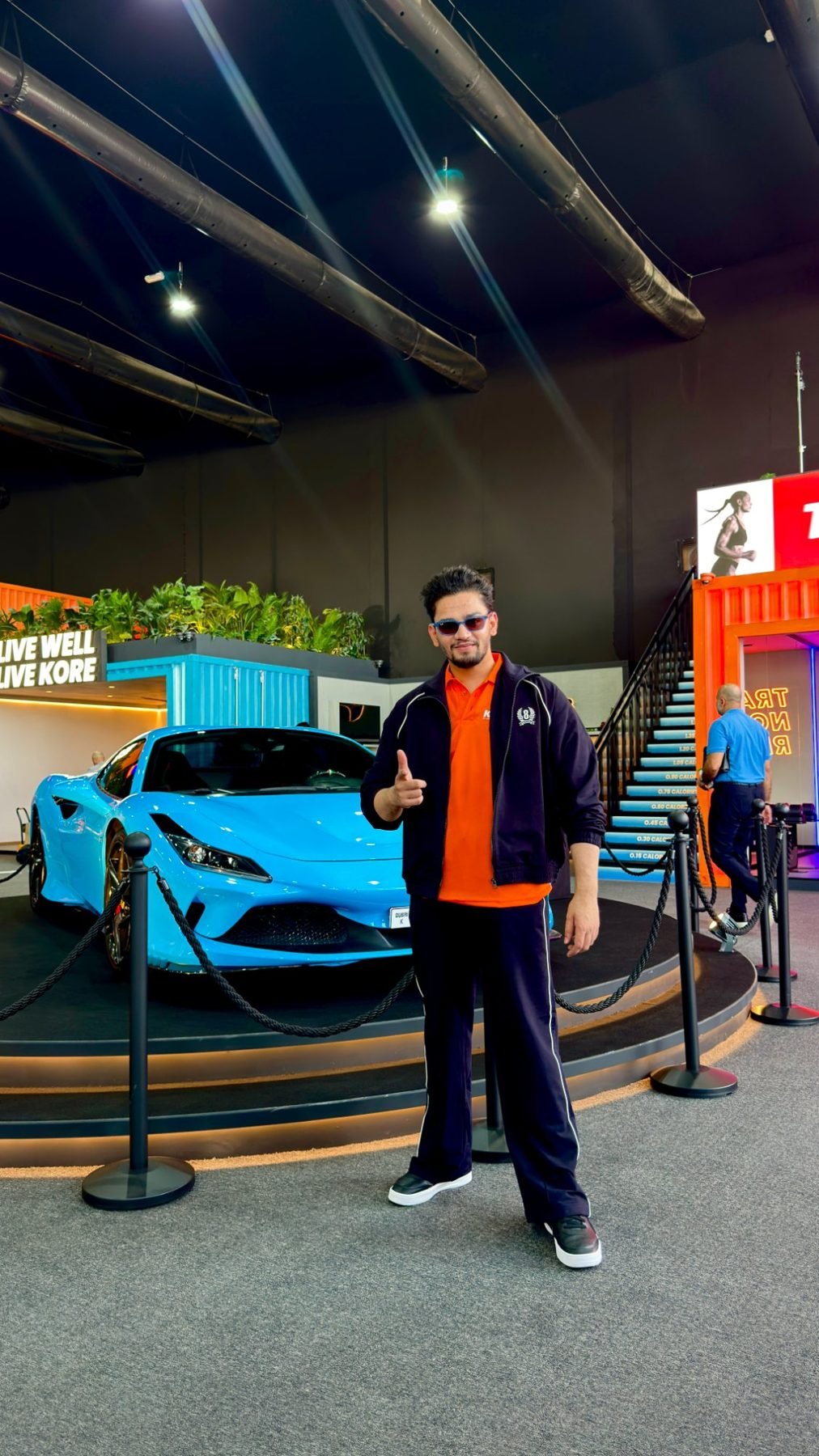 Shah Hussain posing next to a blue Ferrari at the KORE by Imtiaz sneak peek event in Dubai under the Live Well, Live KORE sign